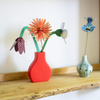 A decorative arrangement of paper flowers in a bright coral-red vase on a wooden shelf, with a smaller blue flower in a pale patterned vase beside it against a white wall.