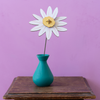 A white daisy flower with a yellow center on a thin stem, displayed in a small teal vase resting on a rustic wooden table against a solid lavender-purple background.