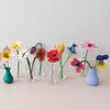 A row of small clear glass vases on a light beige surface against a white wall, each holding a different colorful paper flower or leaf flower arrangement. The vases include teal, lavender, and clear glass, with flowers in yellow, pink, red, blue, purple, and white.