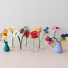A row of small clear glass vases on a light beige surface against a white wall, each holding a different colorful paper flower or leaf flower arrangement. The vases include teal, lavender, and clear glass, with flowers in yellow, pink, red, blue, purple, and white.