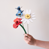 A hand holding three colorful wooden flower cutouts on thin sticks against a plain light background: a white daisy with a yellow center, a red flower, and a blue flower with green stem.