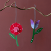 Two colorful hanging flower ornaments shaped like a red rose and a purple tulip with green stems, suspended from a natural tree branch against a solid dusty rose background.