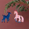 Christmas ornaments shaped like a blue reindeer and a pink horse, hanging from a green pine branch against a solid terracotta background. Made of Cardboard and colorfully decorated with vegetable inks