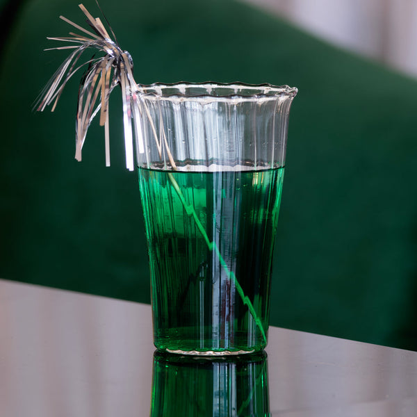 Green cocktail in a glass with a decorative straw on a reflective surface.