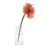 An orange artificial daisy flower with a green stem, displayed in a clear glass vase against a solid white background.