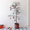 A small potted Christmas tree stands against a white wall on a checkered rug, decorated with colorful hanging ornaments. A red box and a few loose ornaments sit on the floor to the left.
